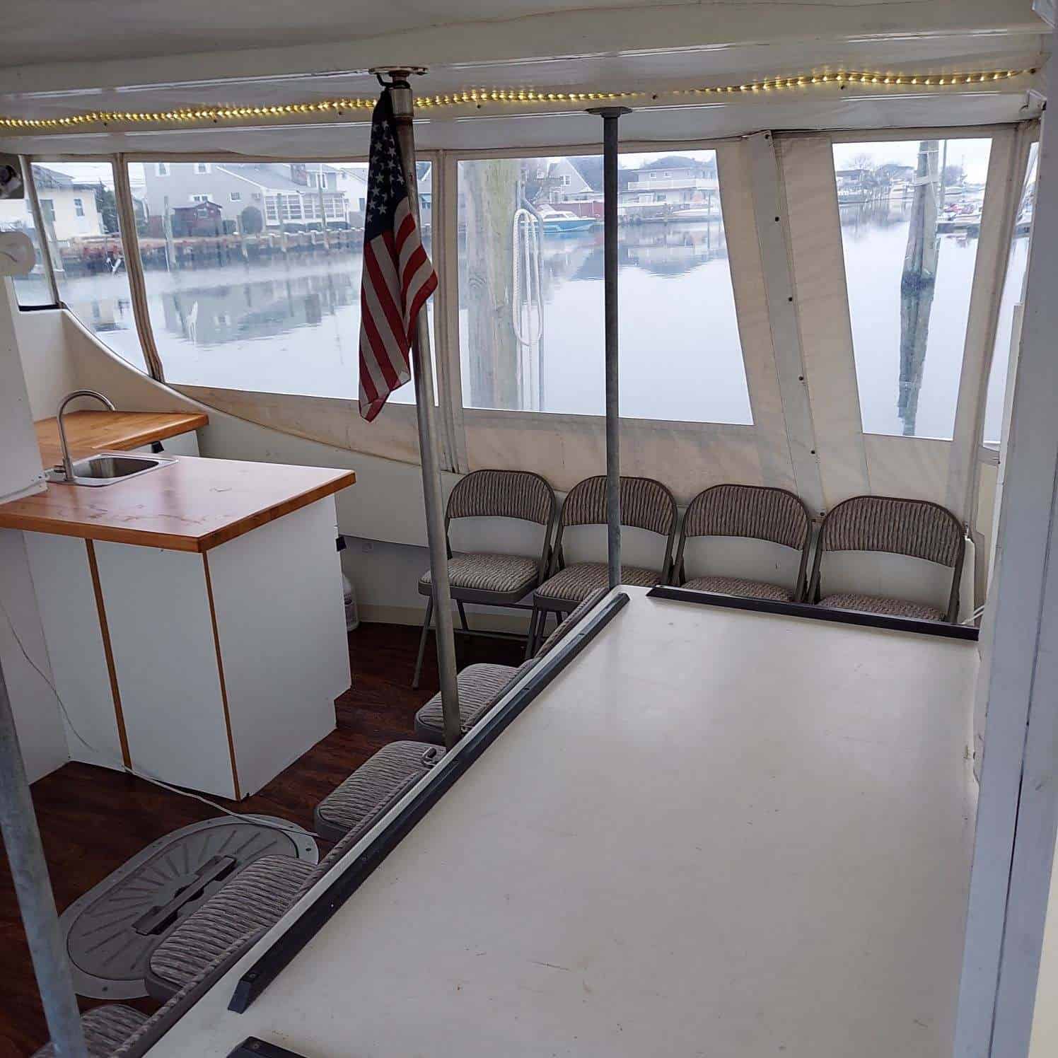 The image shows the interior of a boat with a minimalist design. The space has several gray chairs lined up along the walls, a small kitchenette on the left with a sink and countertop, and an American flag hanging from a pole. Large windows provide views of the marina outside, perfect for an unattended sea burial in Queens County.