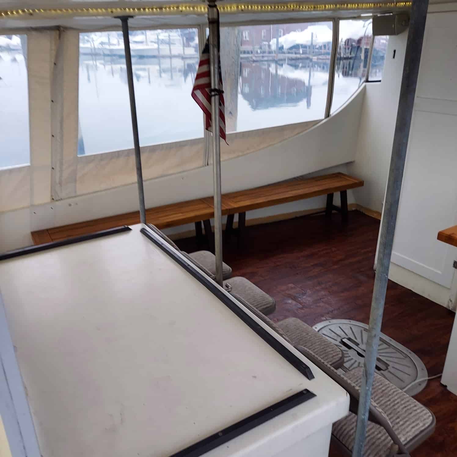 Interior of a boat with white walls and a wooden floor. Benches line the walls, and an American flag is hung near the rear, which is enclosed with clear plastic windows. The view outside shows reflections of boats in calm water, ideal for an unattended sea burial in Nassau County.