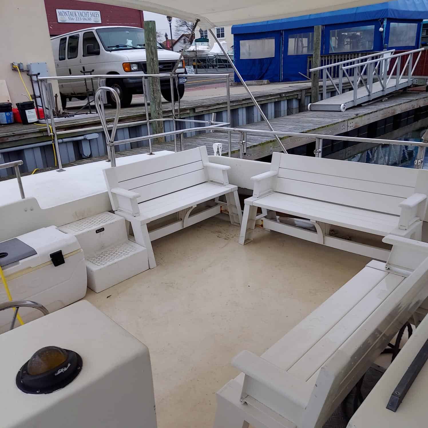 A view of the deck of a boat with white benches arranged in a U-shape under a canopy. The background shows a docked white van and a blue and red building alongside the dock. Various hoses and equipment are seen on the deck, ready for an unattended sea burial in Nassau County.