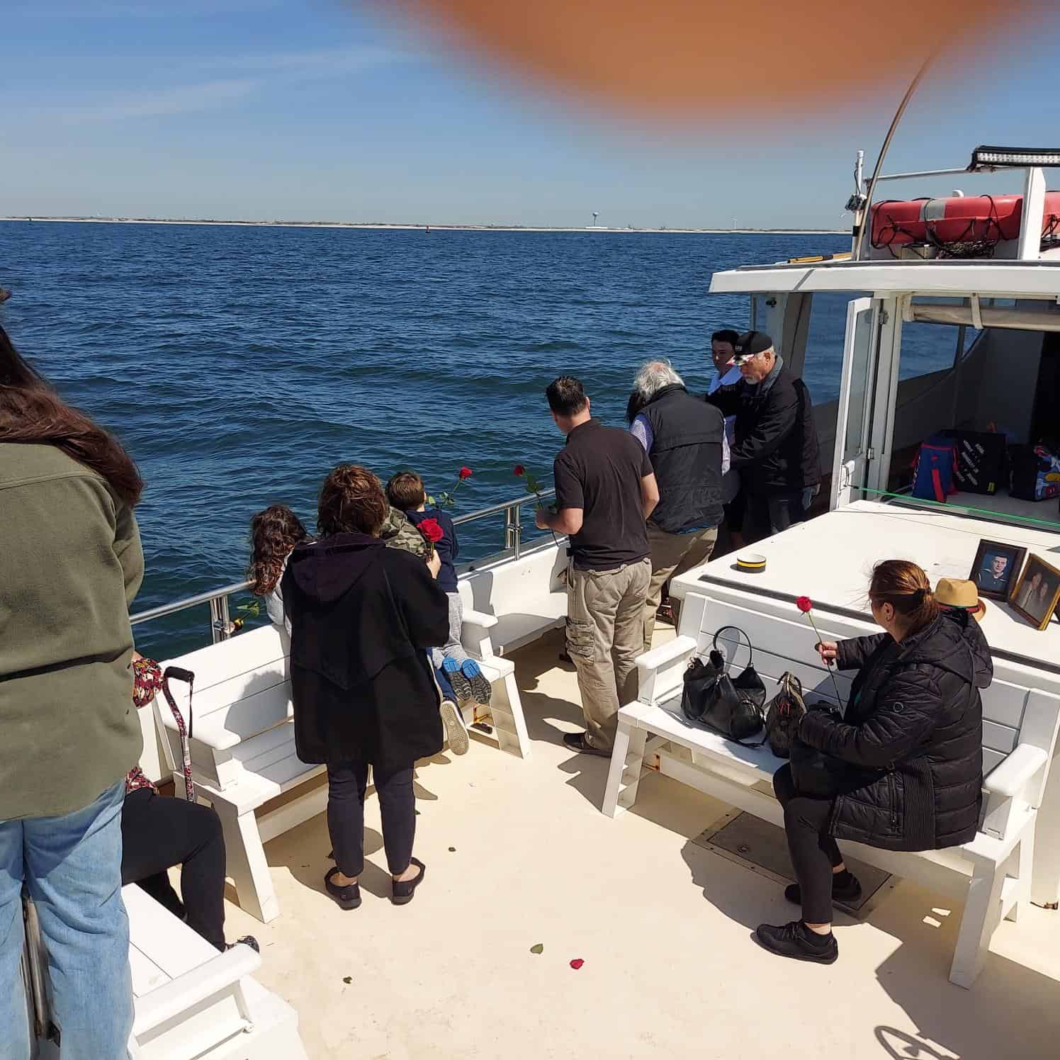 A group of people gathered on a boat off the coast for an unattended sea burial in Nassau County. Some are sitting on benches while others stand, looking out over the water. A few individuals hold or throw flowers into the ocean. One person's finger partially covers the top right corner of the image.