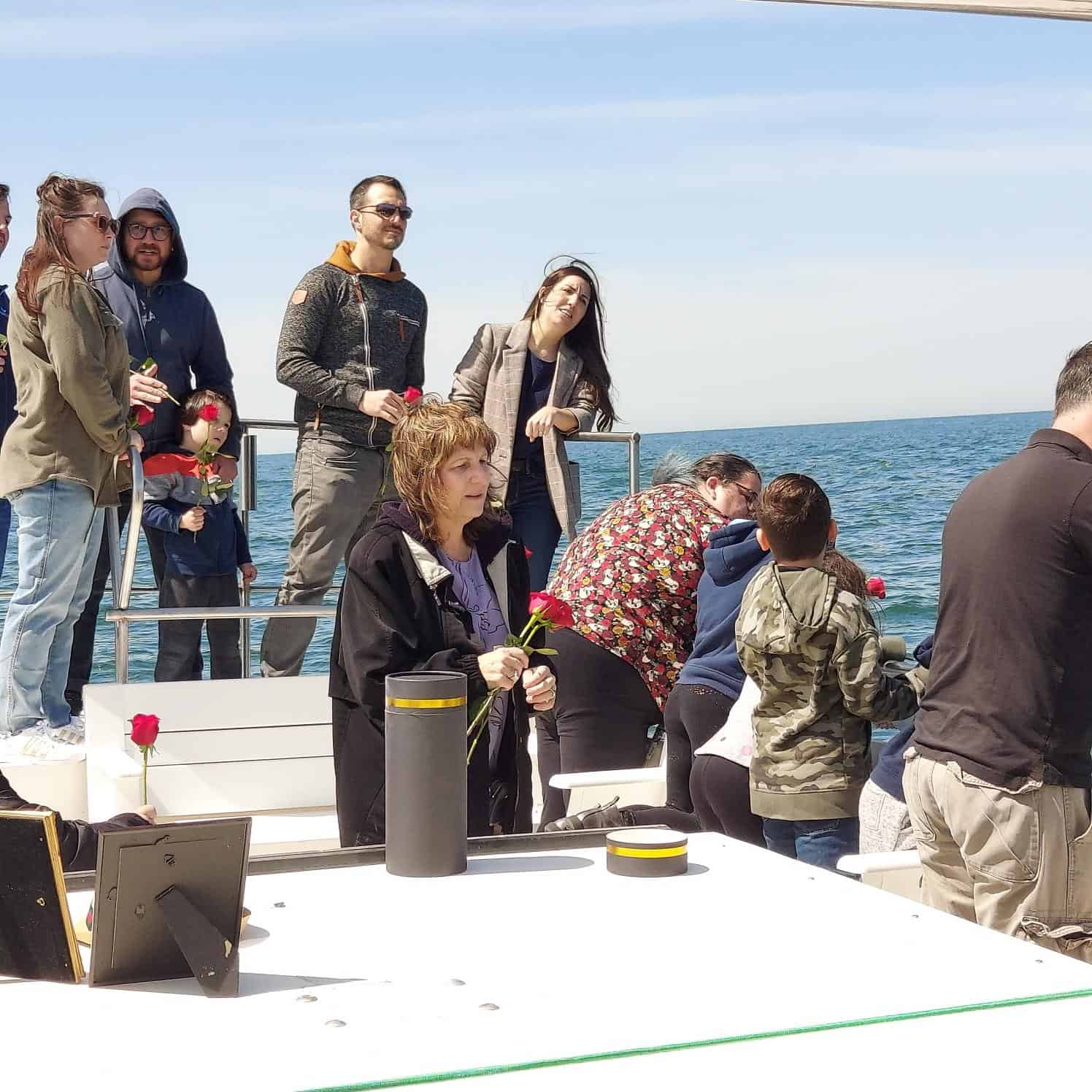 A group of people standing on a boat on a sunny day, holding roses and looking out over the water during an unattended sea burial in Queens County. Some are wearing jackets and sunglasses. There are various objects and containers on a white table in the foreground. The sky is clear and blue.