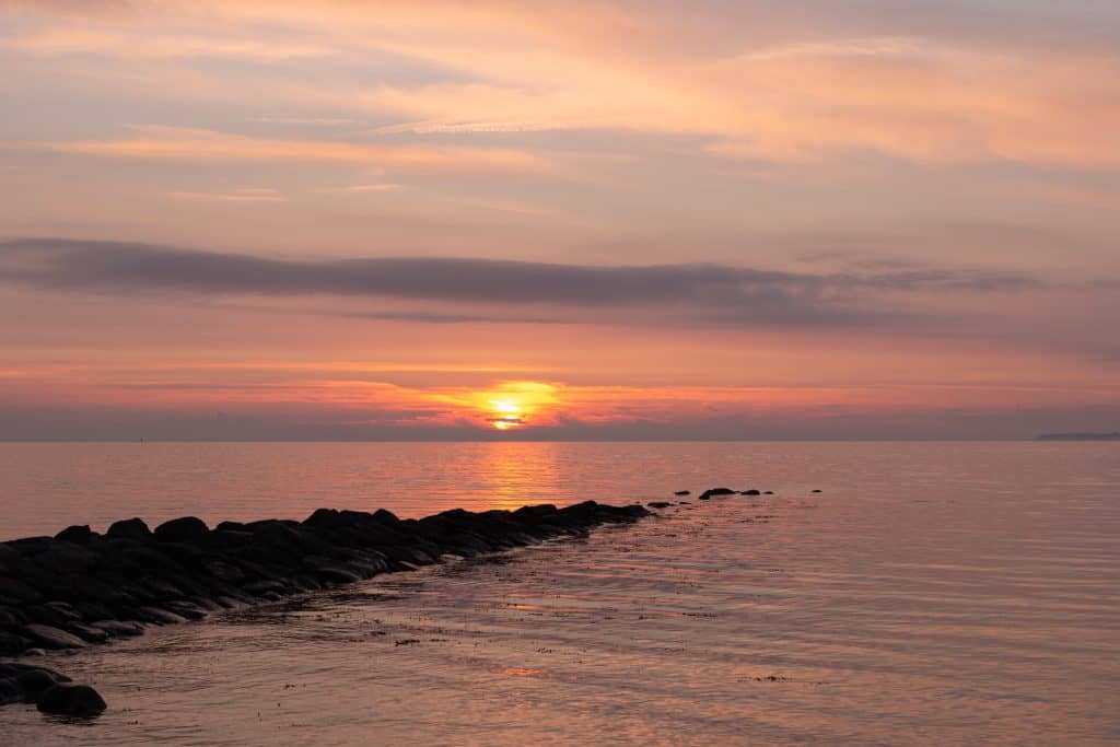 A serene sunset over a calm body of water, with the sun partially hidden by clouds and casting a pink and orange hue across the sky and water. A rocky breakwater extends into the water, creating a peaceful, reflective scene perfect for contemplating or even holding a Pet Sea Burial Nassau County.