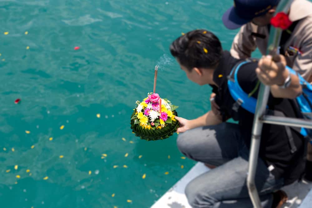 Two people releasing a decorated leaf boat adorned with flowers and incense onto the water, participating in a traditional floating ceremony. They are standing on the edge of a boat in Nassau County, with the clear turquoise water below them—much like an intimate pet sea burial.